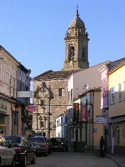 Kirchturm mit Uhr in Melide, Galicien, mit bunten Gebäuden und Autos im Vordergrund.