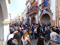 Many people on a street in Palos de la Frontera with colorful buildings and balconies.