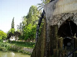 Altes Wasserrad neben einem Fluss mit Bäumen und dem Huerta-Museum im Hintergrund.