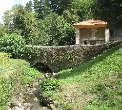 Puente de piedra con ermita y río rodeado de vegetación.
