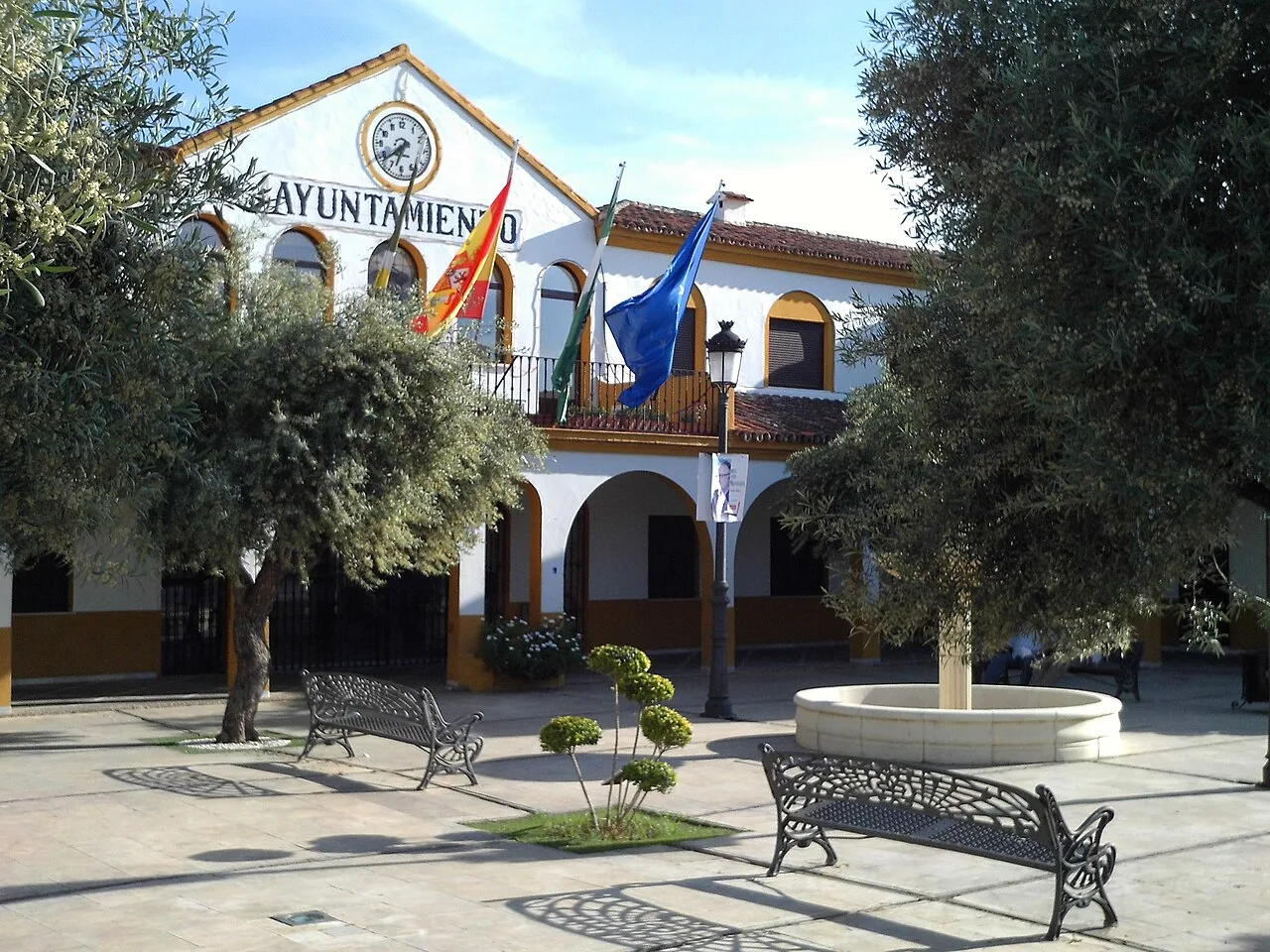 Plaza con árboles, bancos y un edificio con banderas y reloj.
