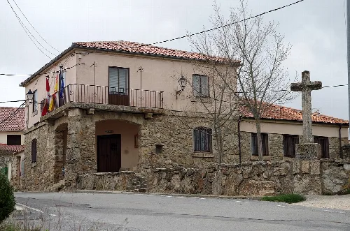 Stone building with balcony and flags, next to a cross in the square.