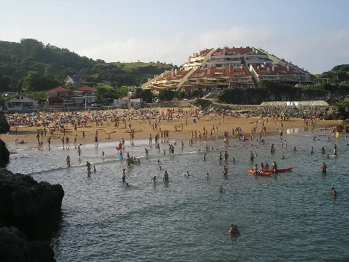 Playa con muchas personas en el agua y un complejo de apartamentos en la colina.