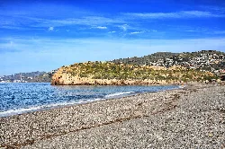 Beach with sand and blue water, rocky promontory and clear sky.