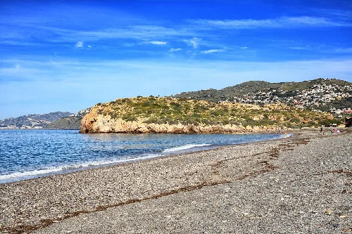 Strand mit Sand und blauem Wasser, felsige Landzunge und klarer Himmel.