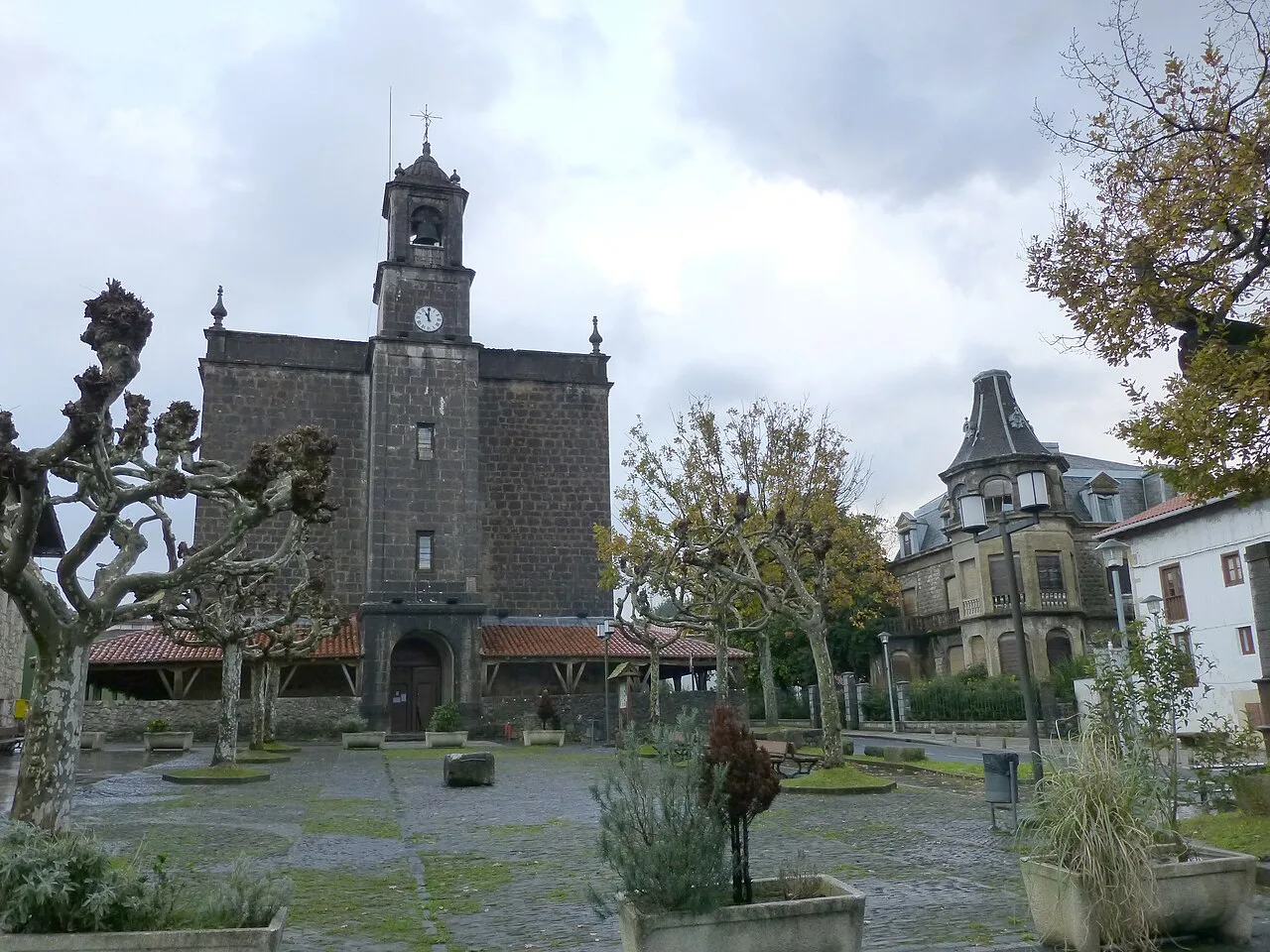 Kirche von Ispáster mit Turm und Uhr, in einem Platz mit geschnittenen Bäumen.