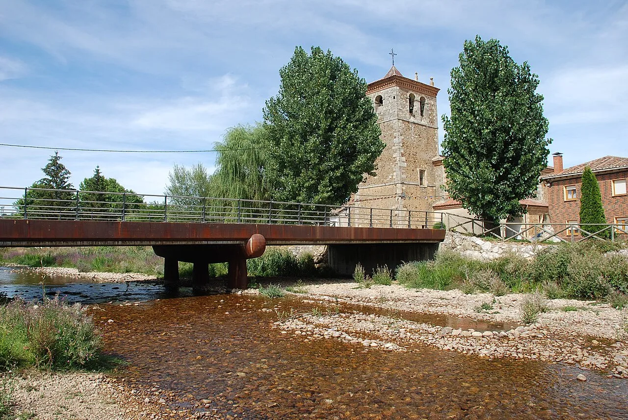Wooden bridge over a river in Báscones de Ojeda with church and trees.