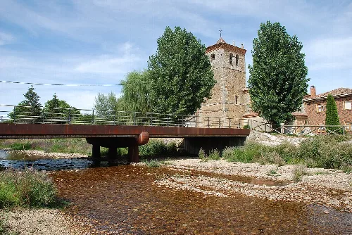 Wooden bridge over a river in Báscones de Ojeda with church and trees.