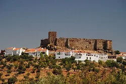 Stone castle on a hill with white houses and trees in the foreground.