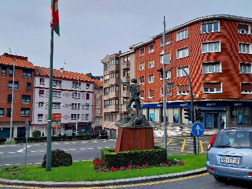 Escultura en una plaza con edificios y coche en Ortuella.