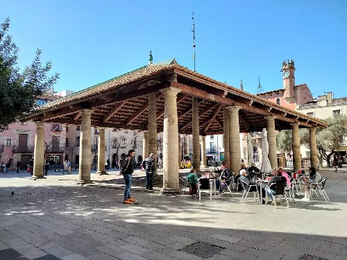 Holzstruktur mit Steinsäulen auf dem Hauptplatz von Granollers, mit sitzenden und gehenden Menschen.