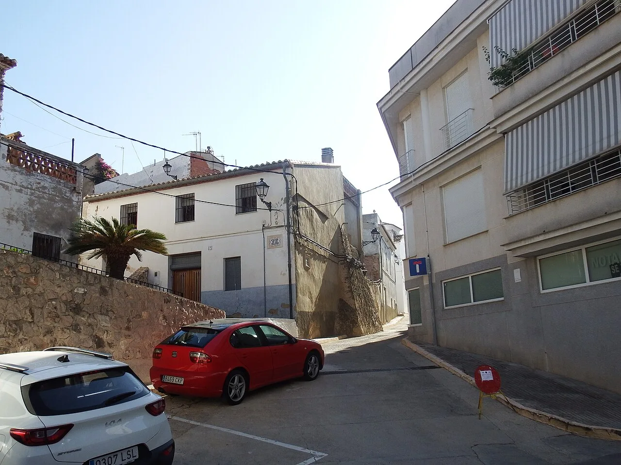 Coches estacionados en una calle de La Font d'en Carròs con edificios y una palmera.