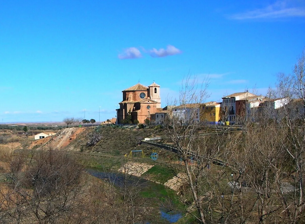 Vista de una iglesia y casas en Vinaceite, en una colina con árboles y un río.