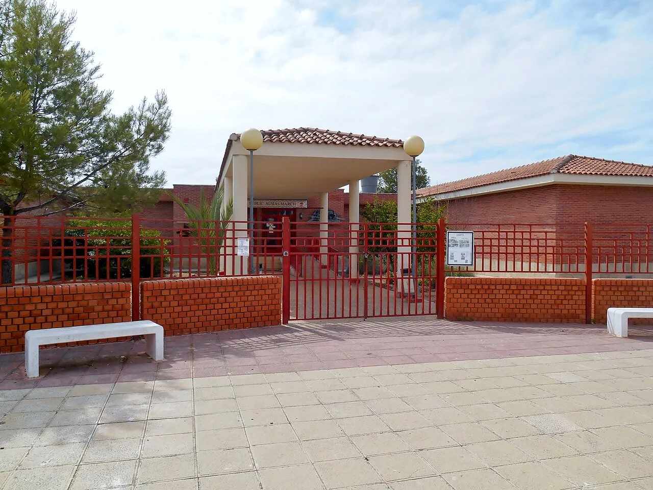 Entrada de una escuela con verja roja y bancos en la plaza.