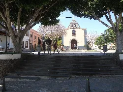 Iglesia en la plaza de Arona, con árboles y personas paseando.