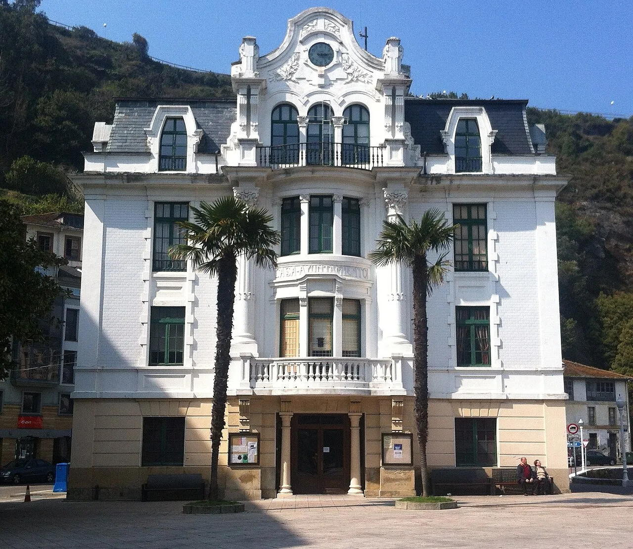White building with large windows, balconies, and two palm trees at the entrance in L.luarca.
