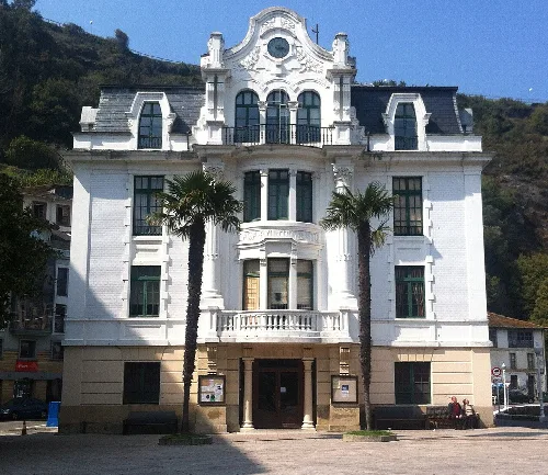 Edificio blanco con ventanas grandes, balcones y dos palmeras en la entrada en L.luarca.