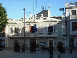 Weißes Gebäude mit Flaggen an der Fassade und Pflanzen auf dem Platz.