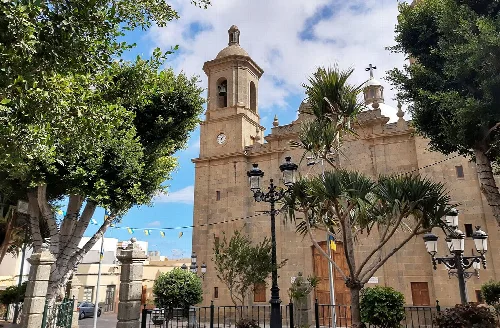 Fassade der Kirche San Sebastián in Agüimes mit Bäumen und Laternen in der Plaza.