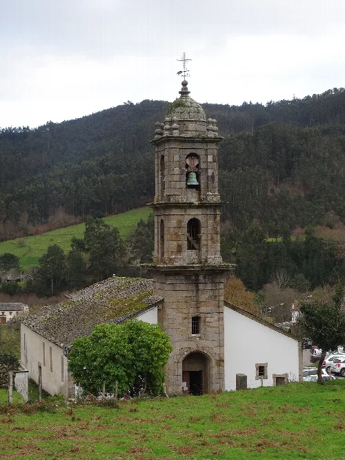 Steinerner Kirchturm mit Glocke und Kreuz auf der Spitze, in einer ländlichen Umgebung mit Hügeln und Bäumen.