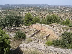Ruinas de una ciudad ibérica en una colina con vegetación y campos en el fondo.