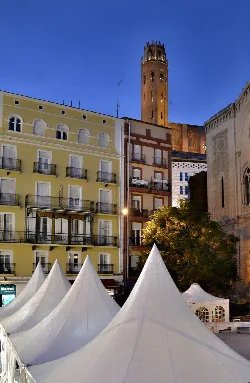 Seu Vella Tower and nearby buildings in Lleida with white tents in the foreground.