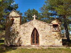 Pequeña ermita de piedra con campanario y cruz en la cima, rodeada de árboles.