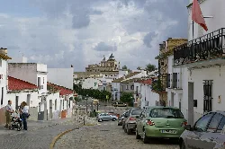 Street view in Santiponce with cars, people, and the monastery in the background.