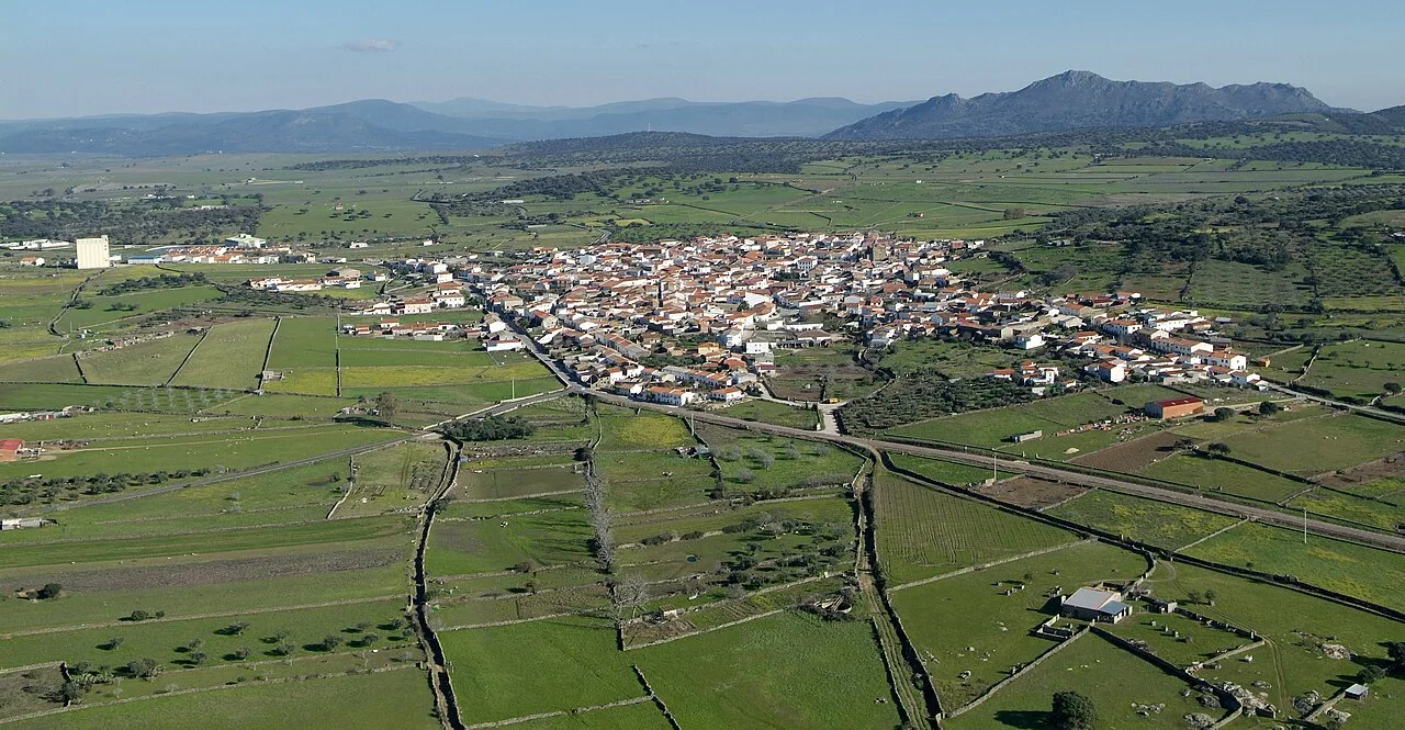 Vista aérea de un pueblo con campos verdes y montañas al fondo.