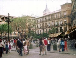 Square with trees, people walking, and historic buildings in Toledo.