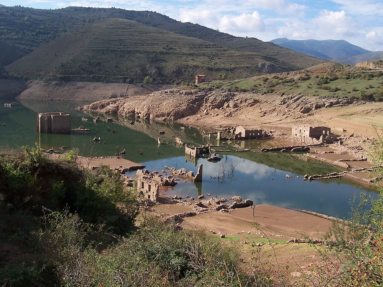 Ruinas de edificios antiguos junto a un embalse en un valle rodeado de montañas.