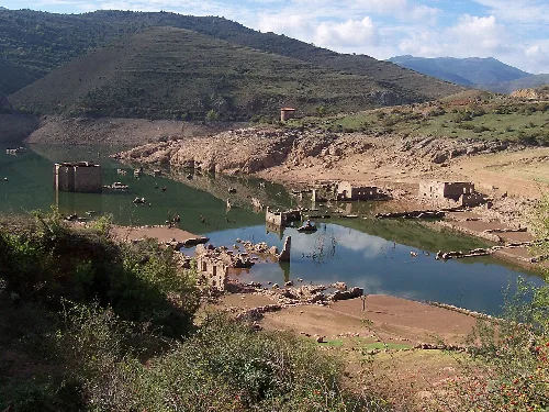 Ruinen alter Gebäude neben einem Stausee in einem Tal mit Bergen im Hintergrund.