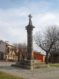 Steinsäule mit Kreuz oben in einem Platz, umgeben von Bäumen und Gebäuden.