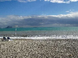 Beach with grey sand, sea, cloudy sky, and two lounge chairs in the foreground.