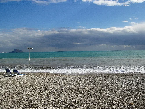 Strand mit grauem Sand, Meer, bewölktem Himmel und zwei Liegestühlen im Vordergrund.
