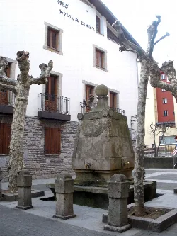 Old fountain with three spouts in an urban square, surrounded by trees and buildings.