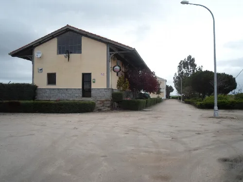 Zweistöckiges Gebäude mit Satteldach und kleinen Fenstern, neben einer Straße und Bäumen.