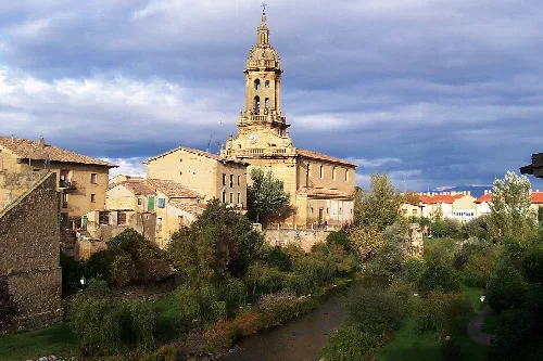 Church and houses in Cuzcurrita de Río Tirón with vegetation and river in foreground.