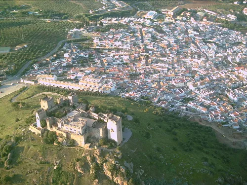 Vista aérea de un castillo medieval en una colina y un pueblo con casas blancas en el fondo.