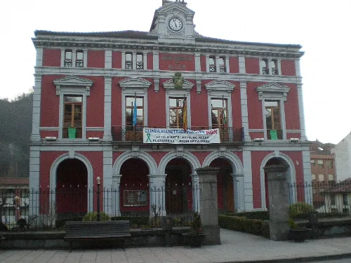 Building with a red facade, clock, and arched entrance in Aller, Asturias.