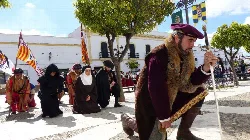 Mann in mittelalterlicher Kleidung kniet in einem Platz mit Mönchen und Flaggen im Hintergrund.
