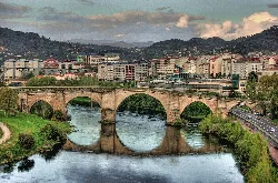 Stone Roman bridge over the river in Ourense, with buildings and mountains in the background.