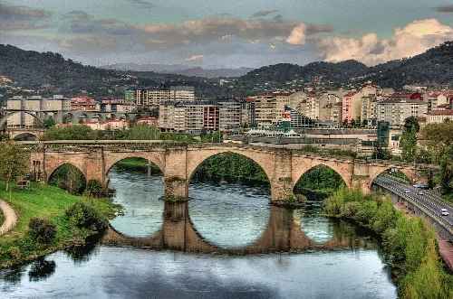 Steinerne römische Brücke über den Fluss in Ourense, mit Gebäuden und Bergen im Hintergrund.