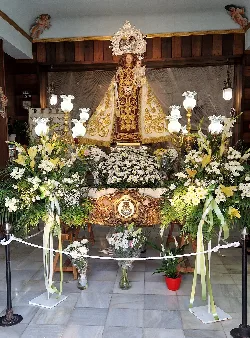 Estatua de la Virgen del Carmen rodeada de flores y velas en una capilla.