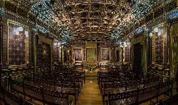 Interior de una sala con asientos de madera y paredes decoradas con cuadros y molduras doradas.