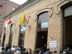 Brick building facade with flags and a crowd of people outside.