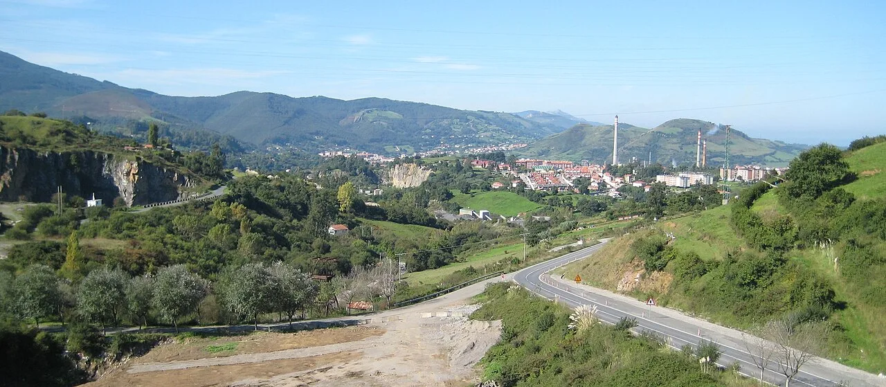 Paisaje con colinas verdes, carretera y vista de pueblos y montañas en el fondo.