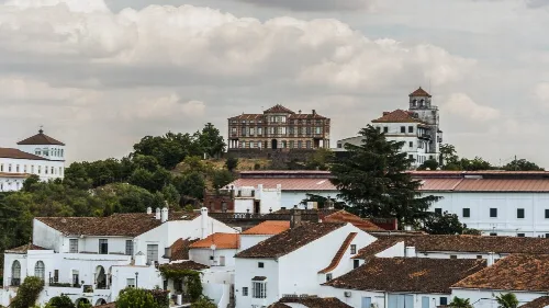 Weiße Häuser und historische Gebäude in Jabugo, Huelva, mit grünen Hügeln und bewölktem Himmel.