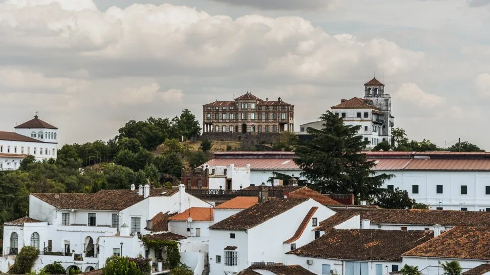 White houses and historic buildings in Jabugo, Huelva, with green hills and cloudy sky.