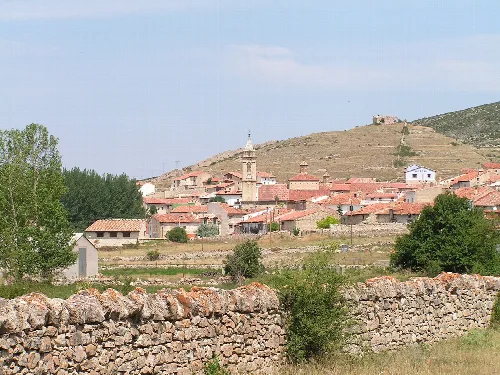 Blick auf ein Dorf mit Häusern und einer Kirche, umgeben von grünen Hügeln und einer Steinmauer im Vordergrund.